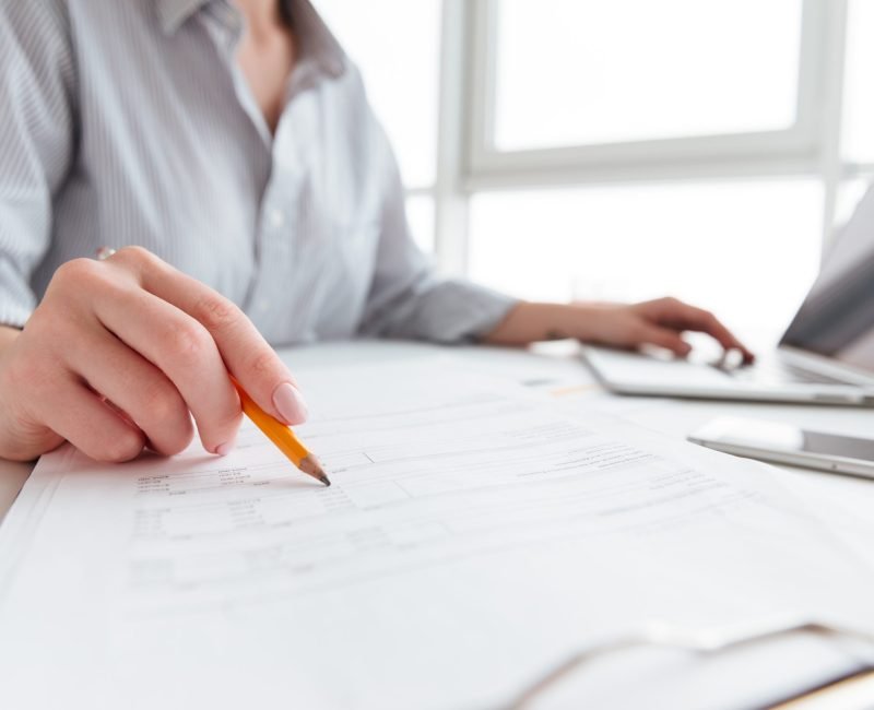 Close up portrait of a woman holding pencil over paper documents while sitting and working with laptop computer at home