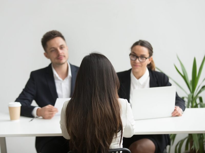 Back close up view of female applicant being interviewed by two HR managers reading her resume, checking data on laptop, asking questions for job position. Employment, hiring, first impression concept